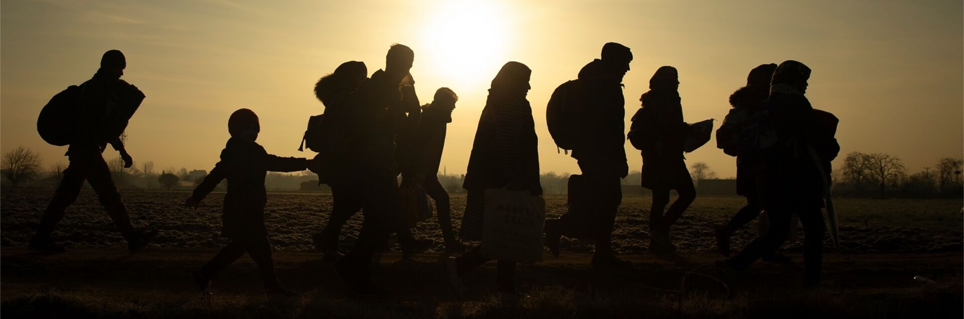 Eine Gruppe von Menschen, darunter Erwachsene und Kinder, geht in Silhouette gegen den Sonnenuntergang. Die Szene vermittelt ein Gefühl von Bewegung und Hoffnung in einer ländlichen Umgebung. (Text KI generiert)