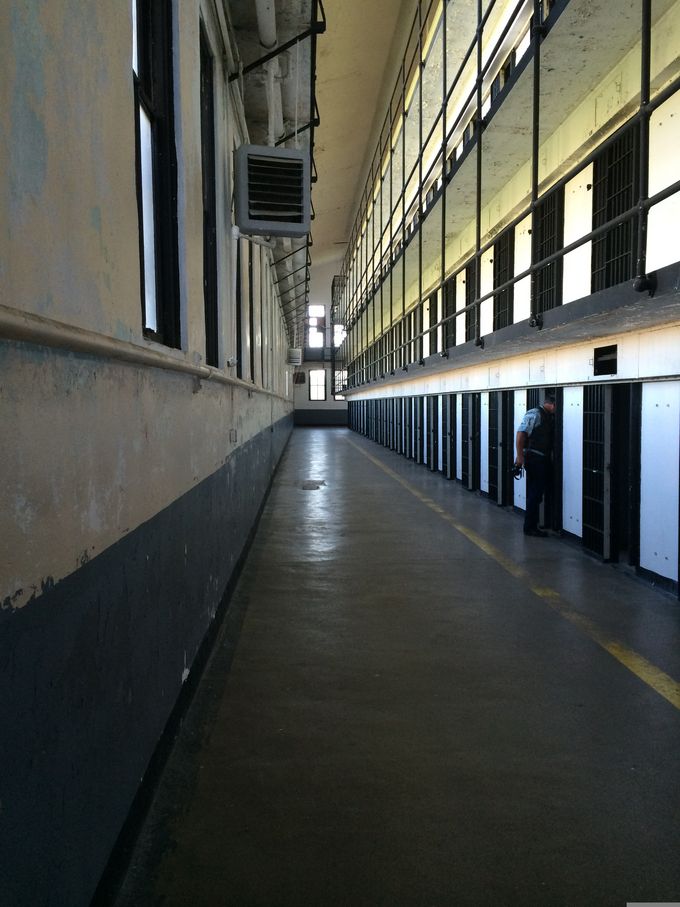 Ein langer, leerer Gang in einem Gefängnis mit hohen Decken und Fenstern. An den Wänden sind Zellen mit geschlossenen Türen. Ein Wächter steht in der Nähe einer Zelle. Der Boden ist grau und der Gang wirkt unheimlich und verlassen. (Text KI generiert)