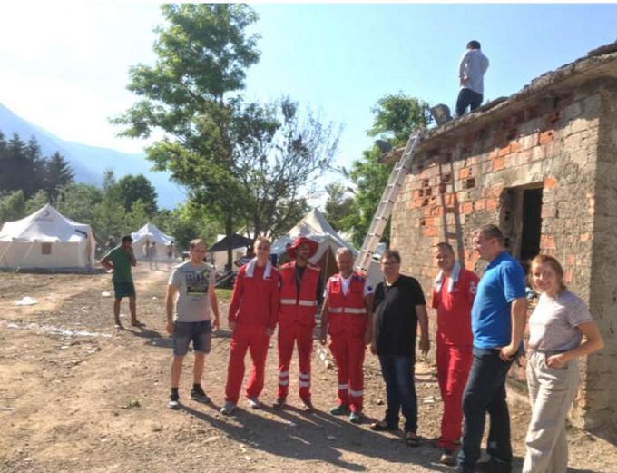 Eine Gruppe von neun Personen steht vor einem alten Gebäude. Einige tragen rote Rettungswesten. Im Hintergrund sind Zelte und Bäume zu sehen. Eine Person arbeitet auf dem Dach des Gebäudes, während eine andere auf einer Leiter steht. (Text KI generiert)
