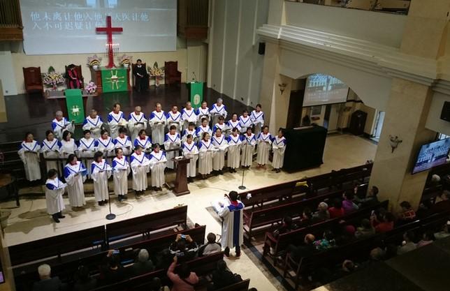 Ein Chor in weißen Roben mit blauen Akzenten steht auf einer Bühne in einer Kirche. Im Hintergrund sind religiöse Symbole und Dekorationen sichtbar. Eine Person spricht vor dem Chor, während das Publikum in den Bänken sitzt und zuhört. (Text KI generiert)