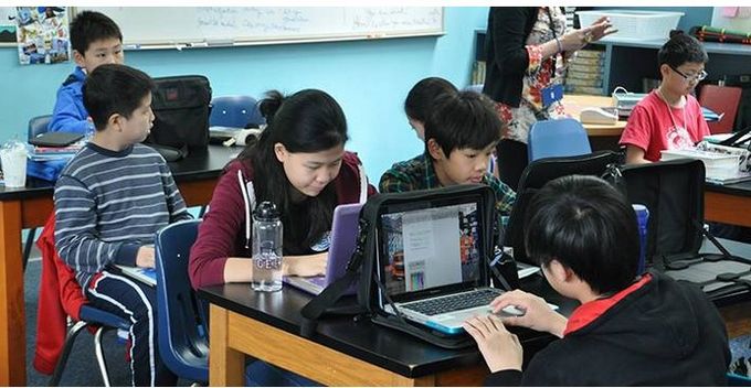 Eine Gruppe von Schülern sitzt in einem Klassenzimmer an Tischen. Einige arbeiten an Laptops, während andere auf ihre Bildschirme schauen. Im Hintergrund ist eine Lehrerin zu sehen, die mit den Schülern spricht. (Text KI generiert)
