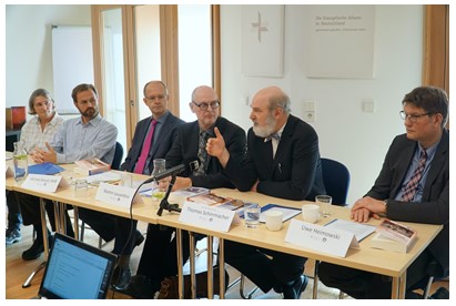Eine Gruppe von fünf Personen sitzt an einem Tisch, der mit Dokumenten und Getränken bedeckt ist. Sie diskutieren, während ein Mann in der Mitte spricht. Im Hintergrund sind Fenster und eine Wand mit einem Kreuz sichtbar. (Text KI generiert)