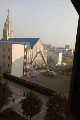 Ein Bagger demoliert ein Gebäude mit einem spitzen Turm. Trümmer liegen auf dem Boden, während die Maschine an der Wand des Gebäudes arbeitet. Im Hintergrund sind weitere Gebäude und ein klarer Himmel zu sehen. (Text KI generiert)